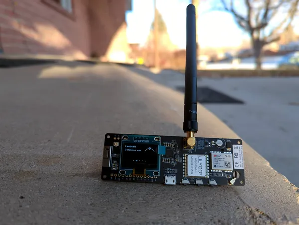 Ein LILYGO T-Beam mit aufgestecktem Display und langer Antenne steht auf einer Mauer.